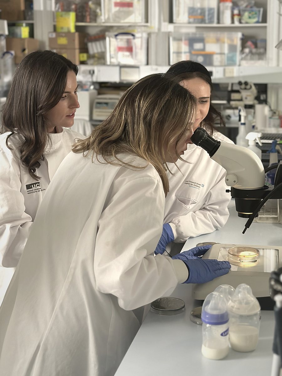 The three BoobyBiome founders in white lab coats at a bench, one looking into a microscope, with bottles of breast milk beside them